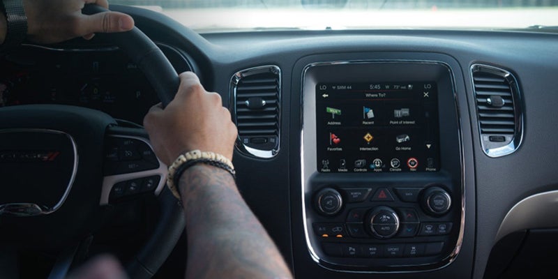 Hands with beaded bracelets on a steering wheel, with a car's infotainment system displaying a navigation menu.