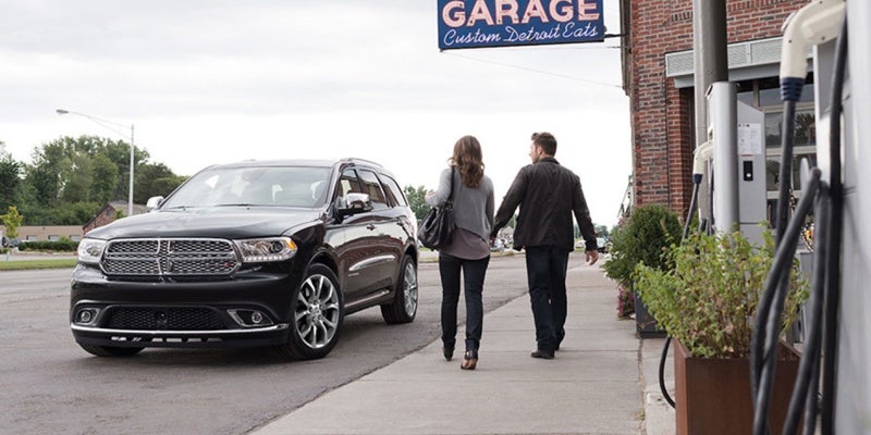 Couple holding hands walks past a black Dodge Durango SUV parked outside of a garage.
