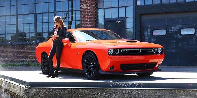 A woman in black leans on an orange Dodge Challenger with black wheels outside a brick and glass building.