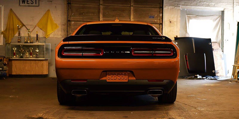 Rear view of a bright orange Dodge Challenger parked in a dim garage with trophies on a shelf to the left.