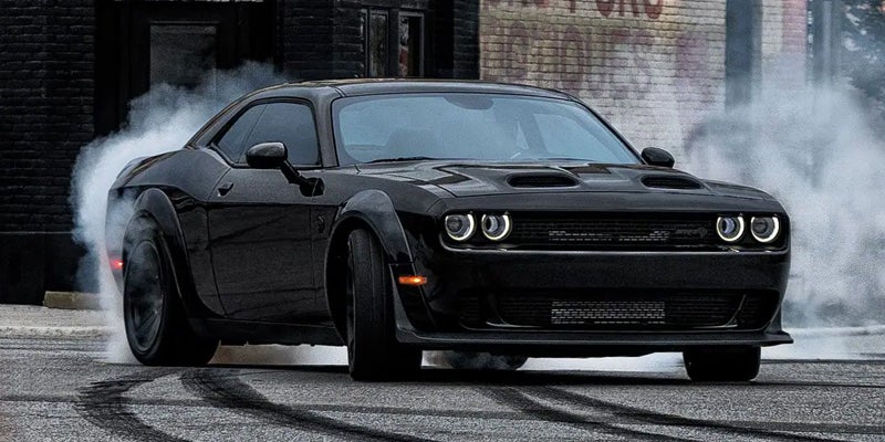 A black Dodge Challenger SRT Demon doing a burnout, with thick white smoke billowing from its tires.