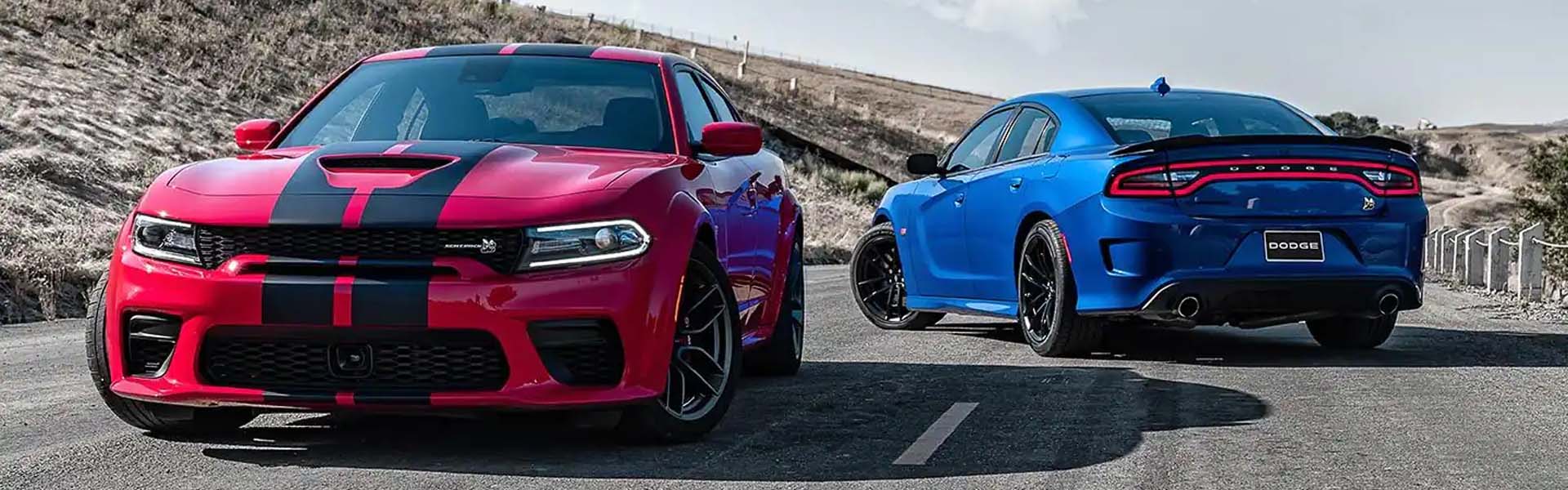 A red Dodge Charger with black racing stripes in front of a blue Dodge Charger, on a paved road.