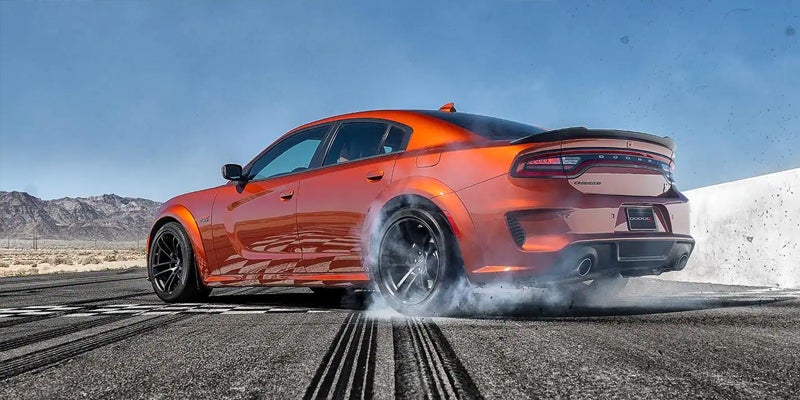 Orange Dodge Charger SRT Widebody performing a burnout, leaving smoke and tire marks on a desert road.