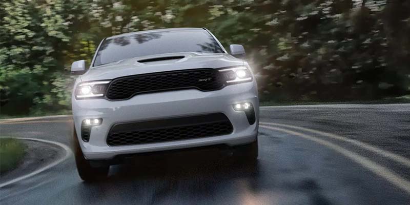 Front view of a white SUV with luminous headlights driving on a wet, winding road with blurred green trees.