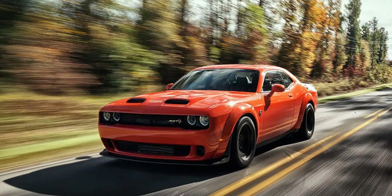 Orange Dodge Challenger SRT Hellcat Widebody drives fast on a tree-lined road with motion blur.
