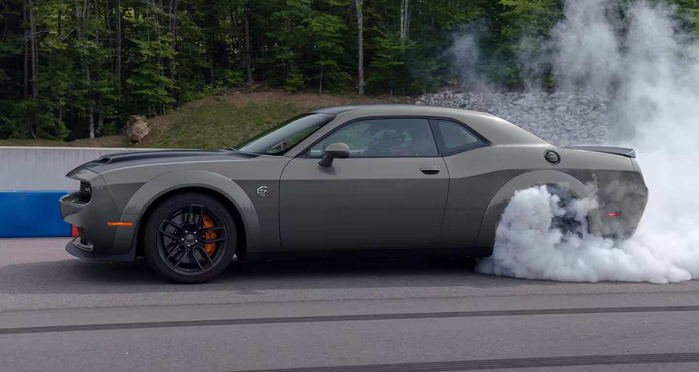 Dark gray Dodge Challenger SRT Hellcat doing a burnout on a track, thick smoke billowing from its rear tires.