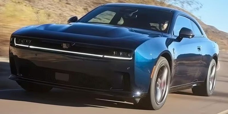A dark blue two-door Dodge Charger sports car driving on a road, with spinning tires and a person visible inside.