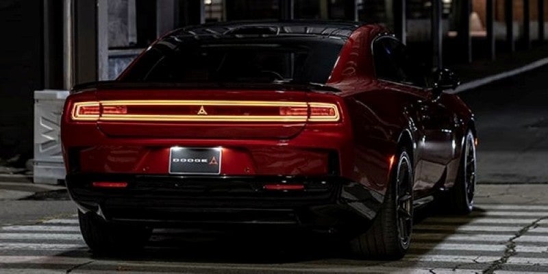 Rear view of a shiny, dark red car with a full-width LED taillight on a dark street, illuminating a Dodge logo.