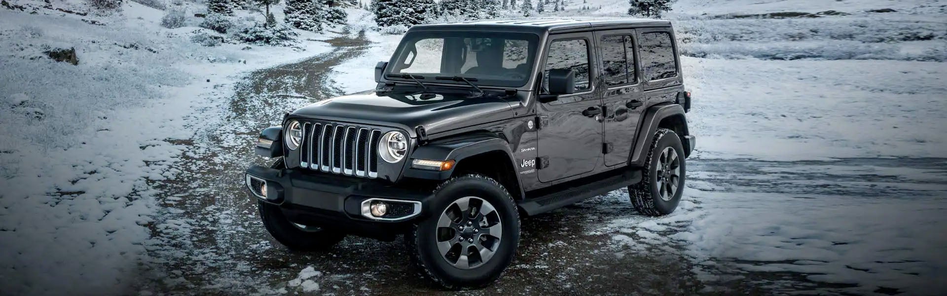 A dark grey Jeep Wrangler parked on a snowy road with pine trees in the background.