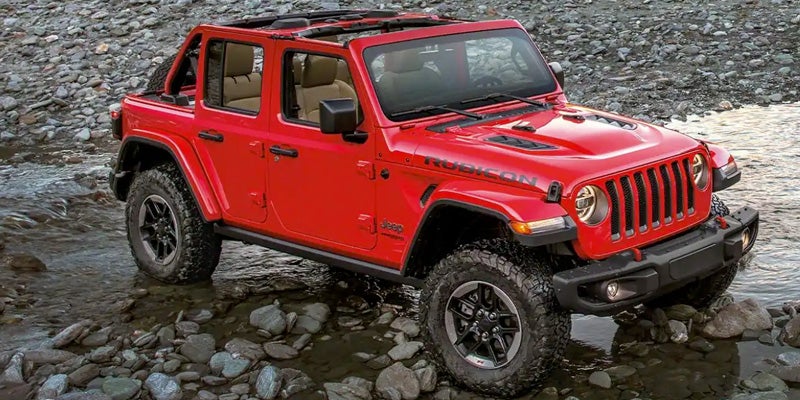 Bright red Jeep Wrangler Rubicon with its top down, left tire in a shallow stream, surrounded by rocks.