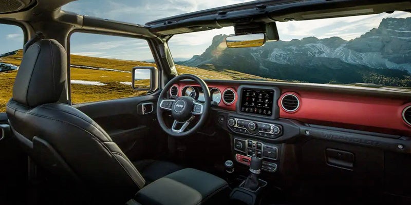 Interior of a black Jeep Wrangler with red dashboard accents, open roof, and mountains in the background.