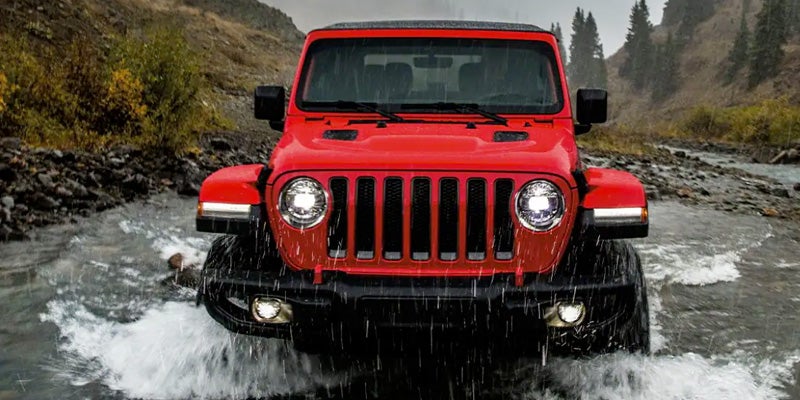 A red Jeep Wrangler Unlimited drives through a rocky stream, its headlights glowing brightly as water splashes around it.