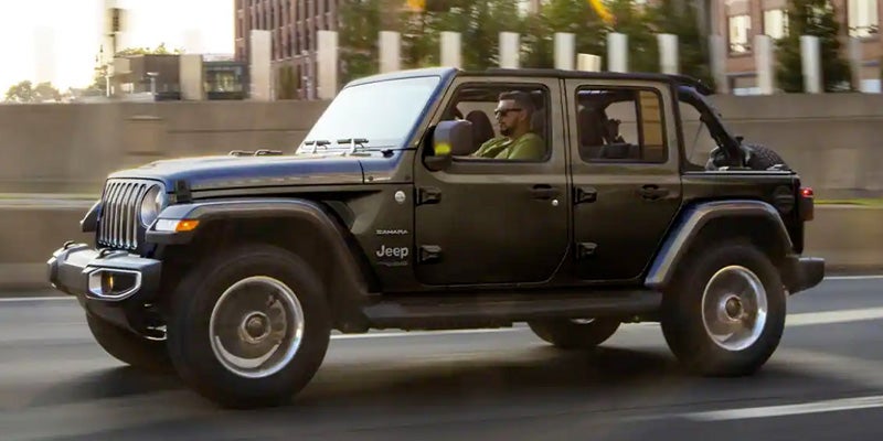 Black Jeep Wrangler Sahara convertible driving left on a highway, with a man wearing sunglasses at the wheel.