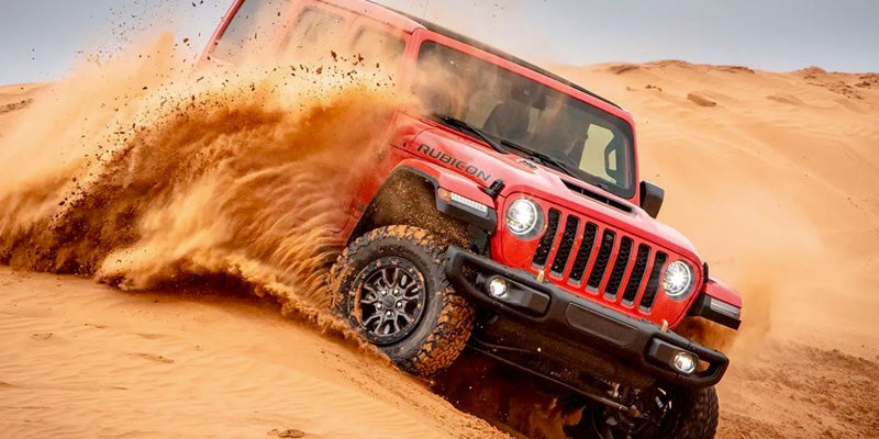 Red Rubicon Jeep kicking up sand while driving off-road up a sandy dune.