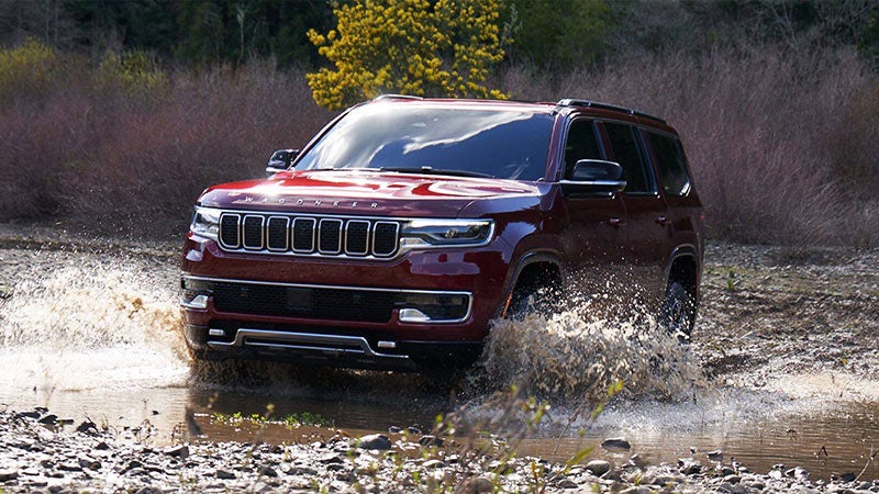 A burgundy Wagoneer SUV drives through a shallow stream, splashing muddy water, with dark bushes in the background.