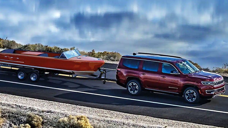 A maroon Wagoneer SUV towing a gleaming orange wooden boat on a trailer, driving right on a two-lane highway.