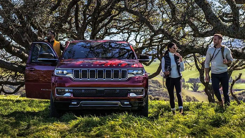 Red Jeep Wagoneer parked under gnarled tree branches, with two hikers to the right and one by the open door.