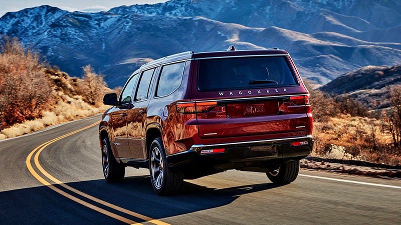 Rear view of a red Wagoneer SUV driving away on a winding road with snow-capped mountains in the background.