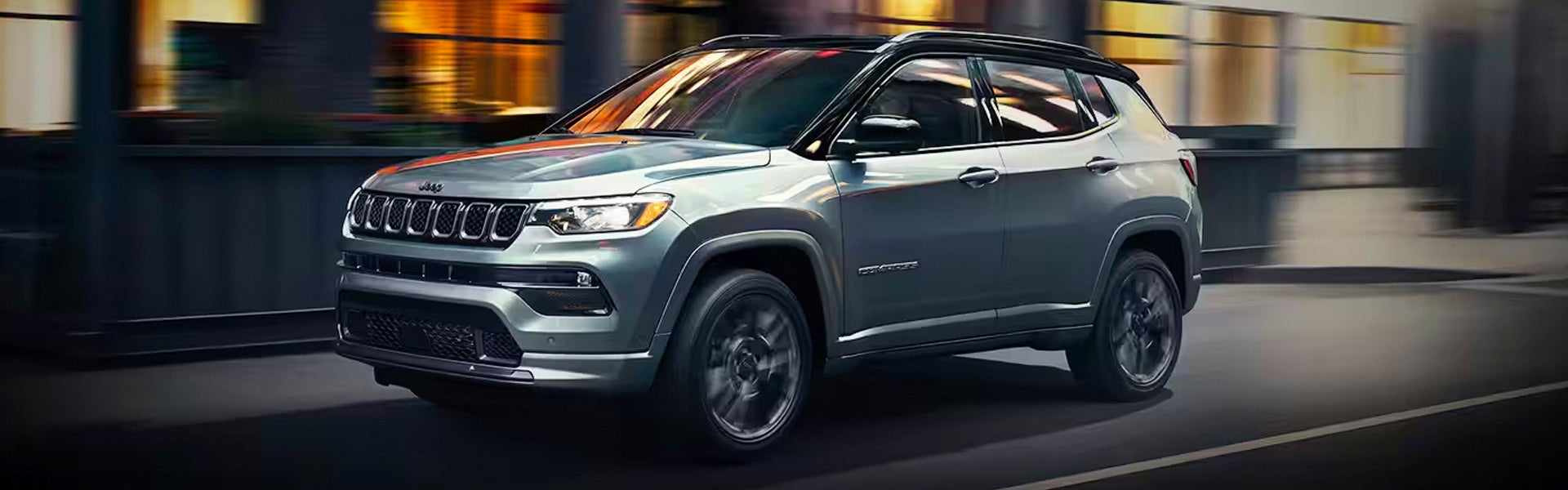 A silver Jeep Compass, blurred with motion, drives forward at night with city lights reflecting on its side.