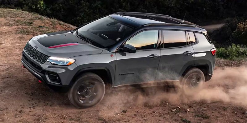 A dark gray Jeep Compass SUV with a black roof and red stripes on the hood drives on a dusty dirt road.