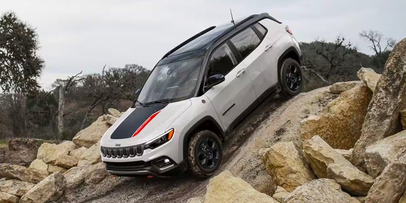 White Jeep Compass Trailhawk with a black hood stripe and red accent drives up a steep, rocky dirt hill.
