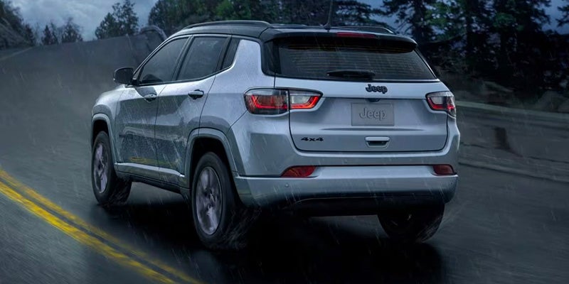 Rear view of a silver 2022 Jeep Compass SUV driving on a winding paved road through a forest.