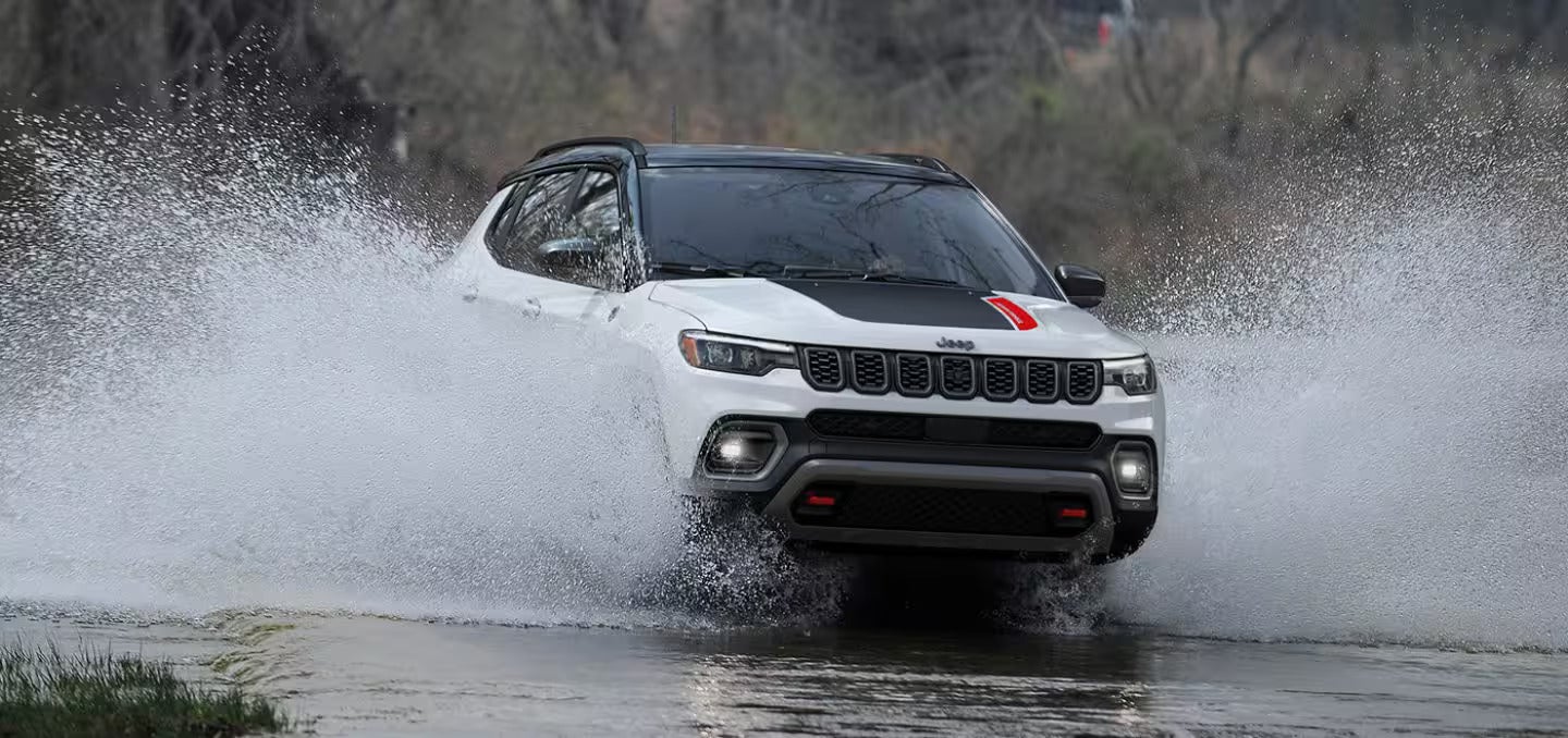 A white Jeep Compass with a black hood drives through a splash of water, moving towards the viewer.