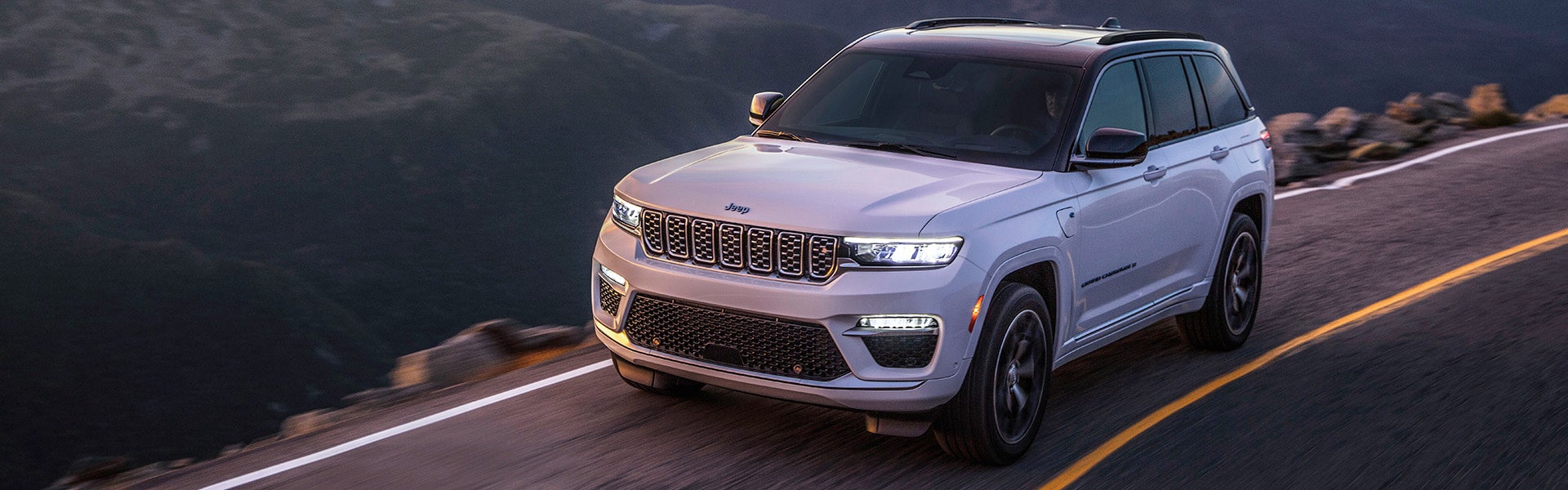 A light grey Jeep Grand Cherokee drives along a winding road with mountains and dark skies in the background.