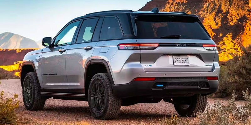A silver and black Jeep Grand Cherokee 4xe parked on a dirt path with brush and mountains in the background.
