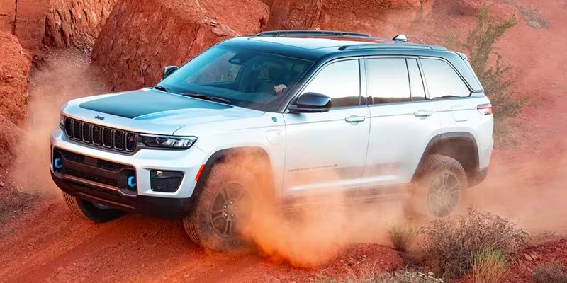 A white Jeep Grand Cherokee 4xe drives through a dusty, rocky, red desert landscape, kicking up orange dust.
