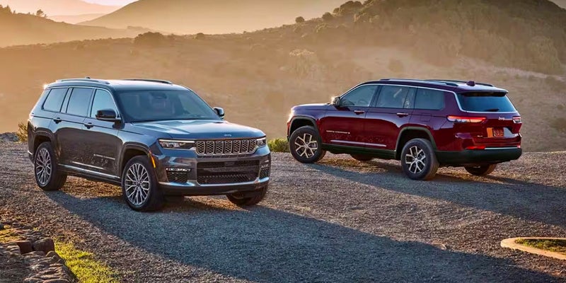 Two Jeep Grand Cherokee Ls, one dark gray and one deep red, parked on a dirt road at sunset.