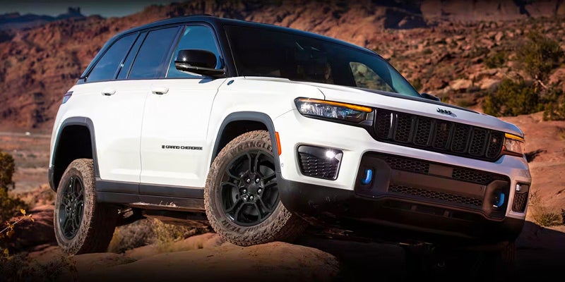 A white Jeep Grand Cherokee, with a black roof and black off-road tires, drives over rocky terrain.
