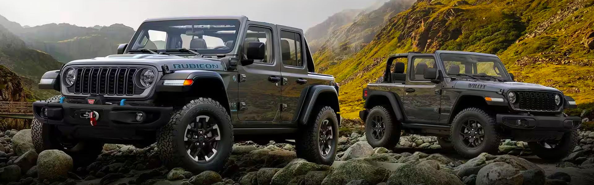 Two dark grey Jeep Wranglers are parked on rocks, one a four-door Rubicon and the other a two-door Willys.