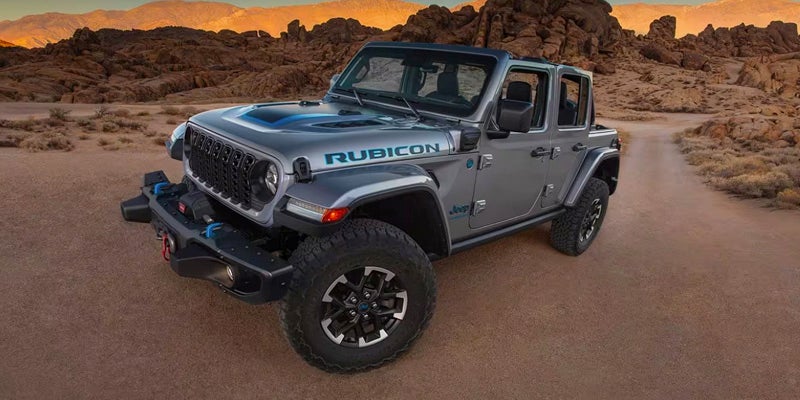 A silver Jeep Wrangler Rubicon 4xe with a blue hood decal sits on a dirt road in a desert landscape.