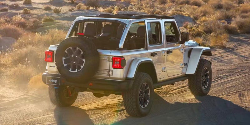 Silver Jeep Wrangler with top up, driving away on a dirt road in a desert landscape.