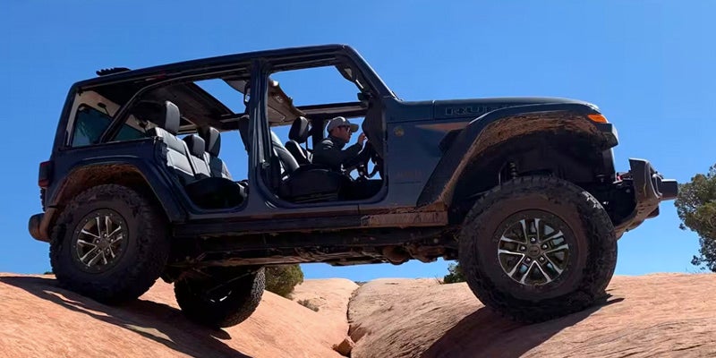 Dark gray Jeep Wrangler Rubicon 4xe with doors removed, off-roading with one wheel in the air.