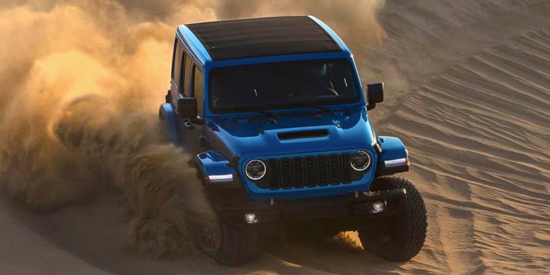 A blue Jeep Wrangler kicking up a large cloud of dust while driving across sand dunes.