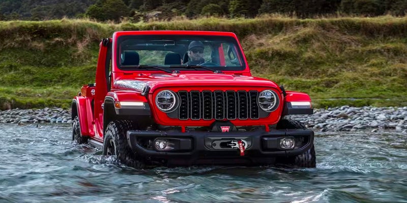 A bright red Jeep Wrangler Rubicon navigating through a shallow river, with a green, rocky bank in the background.