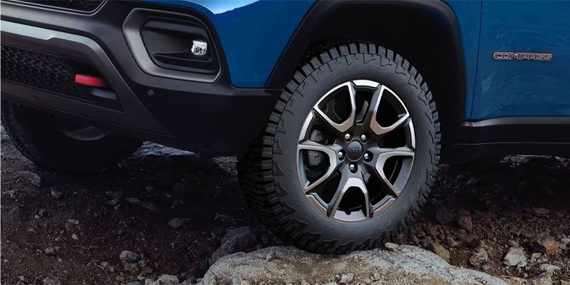 Close-up of a blue Jeep Compass on a rocky, dark ground, showing its rugged tire and front bumper.