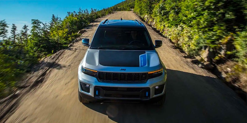A silver Jeep SUV with a black textured hood drives on a narrow dirt road surrounded by green trees on hills.