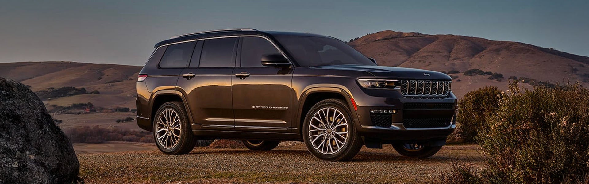 A dark gray Jeep Grand Cherokee L parked on a dirt hill with brown mountains and a blue sky in the background.