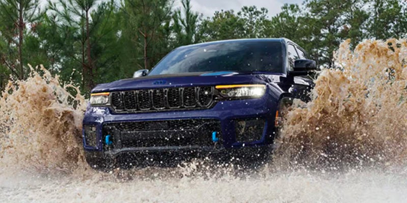 Front view of a dark blue Jeep with bright headlights driving through a large splash of muddy water.