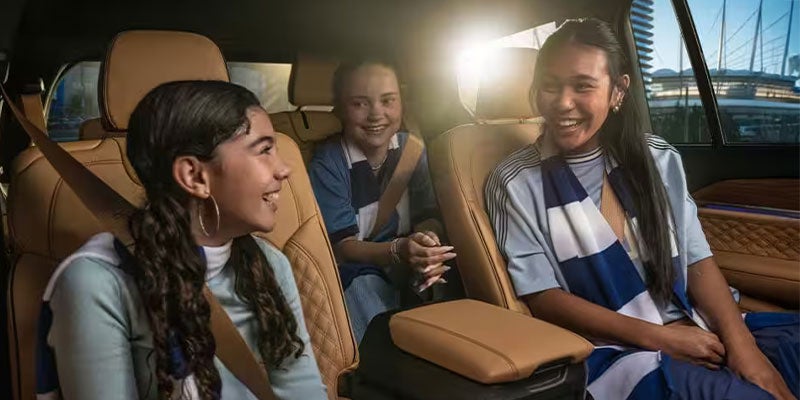 Three joyful girls, possibly sisters, in sports scarves, laughing together in the backseat of a luxurious car.