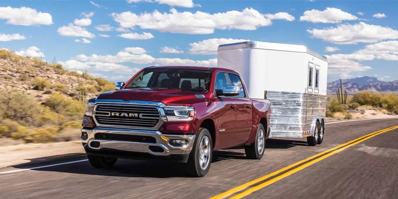 A red Ram pickup truck pulls a white aluminum horse trailer down a desert highway under a blue sky with white clouds