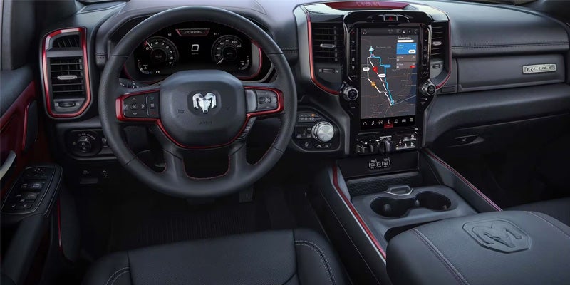 Interior of a Ram truck, showing a black leather steering wheel with red trim, a large vertical screen with a map, and black leather seats with white stitching.