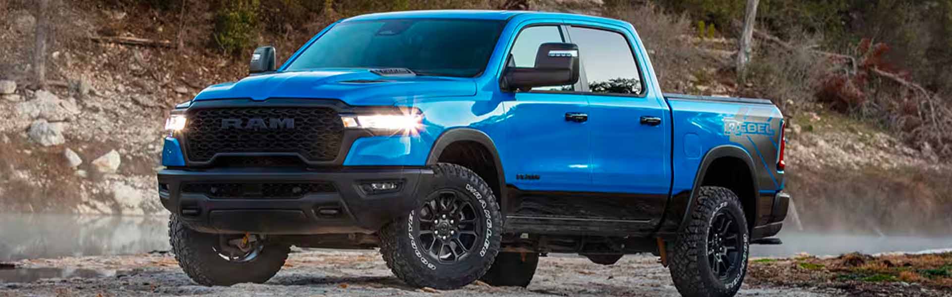 A bright blue Ram Rebel pickup truck with glowing headlights and large off-road tires parked next to a body of water.