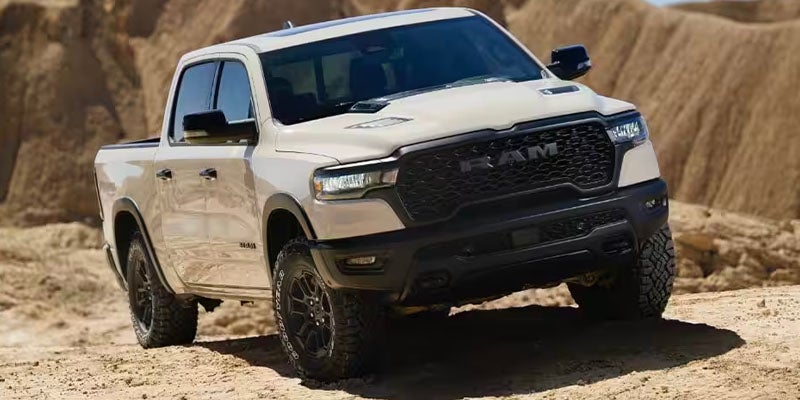 White Ram pickup truck with black grille and wheels, angled on a sandy, uneven terrain with hills in the background.