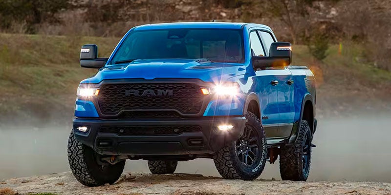 A blue Ram 1500 TRX pickup truck with its headlights on, driving on a dirt road.